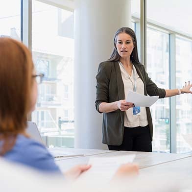 A%20female%20Director%20of%20Nursing%20stands%20in%20front%20of%20a%20conference%20table%20and%20is%20looking%20at%20a%20group%20of%20seated%20employees%20while%20giving%20a%20presentation%20and%20pointing%20to%20some%20information%20on%20a%20whiteboard%20behind%20her.