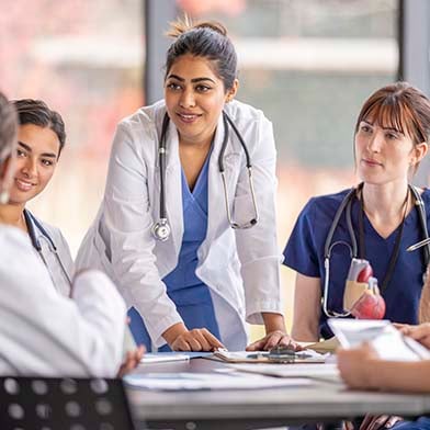 A%20group%20of%20attentive%20nurses%20in%20scrubs%20and%20white%20coats%20gather%20around%20a%20table%20for%20a%20team%20briefing,%20led%20by%20a%20nurse%20standing%20up,%20indicating%20a%20collaborative%20medical%20discussion.