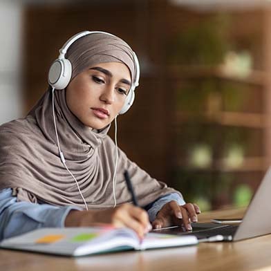 A%20focused%20nursing%20student%20in%20a%20hijab%20is%20studying%20for%20the%20NCLEX%20exam%20at%20a%20desk.%20She%20is%20wearing%20headphones%20and%20looking%20intently%20at%20her%20laptop%20screen%20while%20taking%20notes.%20A%20coffee%20cup%20and%20various%20study%20materials%20are%20organized%20around%20her,%20suggesting%20a%20productive%20study%20session%20in%20a%20quiet,%20indoor%20environment.