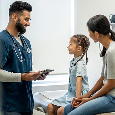 A%20cute%20seven%20year%20old%20girl%20is%20at%20a%20check-up%20appointment%20with%20her%20healthcare%20provider.%20She%20is%20sitting%20on%20the%20examination%20table%20with%20her%20mother.%20She%20is%20looking%20at%20her%20male%20nurse%20practitioner%20and%20smiling%20while%20her%20mother%20is%20seated%20next%20to%20her.