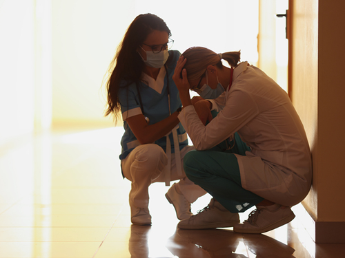 A nurse in a white lab jacket is crouched down and leaning against a wall in a hospital hallway. They have their head down and appear distressed. A second nurse is squatting next to them and offering support. Both are wearing surgical masks, and the hallway is dimly lit.