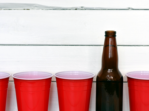 A row of red plastic cups and one amber glass bottle are against a backdrop of the white siding of a building.