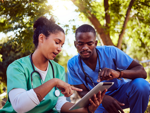 Two nurses are seated outdoors and looking at data on an electronic tablet. They are engaged in conversation as one nurse points to something on the screen.