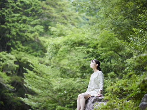 A smiling woman wearing all white is seated in the middle of a lush green forest. 