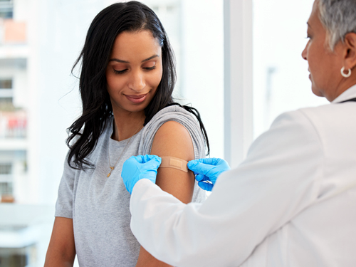A nurse wearing a white lab jacket and blue gloves is placing a Band-Aid on the arm of a seated young woman who has just received her vaccination.