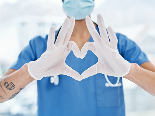 A male nurse wearing a blue scrub outfit and a surgical mask, and gloves is making a heart symbol with his hands held out in front of him. His face is not visible.