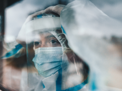 A nurse in full protective PPE is looking out through a glass door of an isolation room. They appear lost in thought.