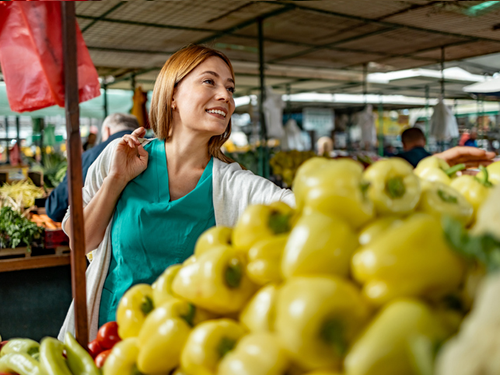 A smiling nurse wearing green scrubs is shopping at a farmers market. She is reaching forward to get some produce. There is a stack of yellow peppers in the foregraound.