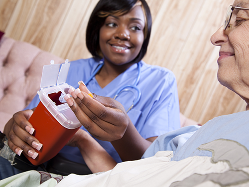 A nurse is seated next to an elderly patient, and she is instructing the patient on how to place a used fingerstick needle into a biohazard sharps container by doing a demonstration. Both are smiling.