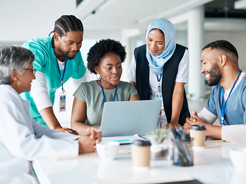 A group of multicultural health care workers are seated and standing near the end of a large table. A laptop is open on the table, and together the group is looking at information on the screen.