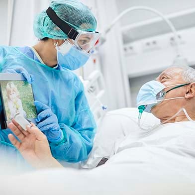 Two nurses are wearing isolation gowns, goggles, and facemasks, and are standing at either side of a patient’s bed. An elderly male patient is in the bed. He is also wearing a mask, and he is looking at a tablet screen the nurse is holding so he can talk with his family over the internet.