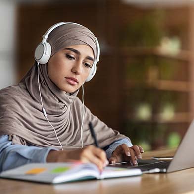 A focused nursing student in a hijab is studying for the NCLEX exam at a desk. She is wearing headphones and looking intently at her laptop screen while taking notes. A coffee cup and various study materials are organized around her, suggesting a productive study session in a quiet, indoor environment.