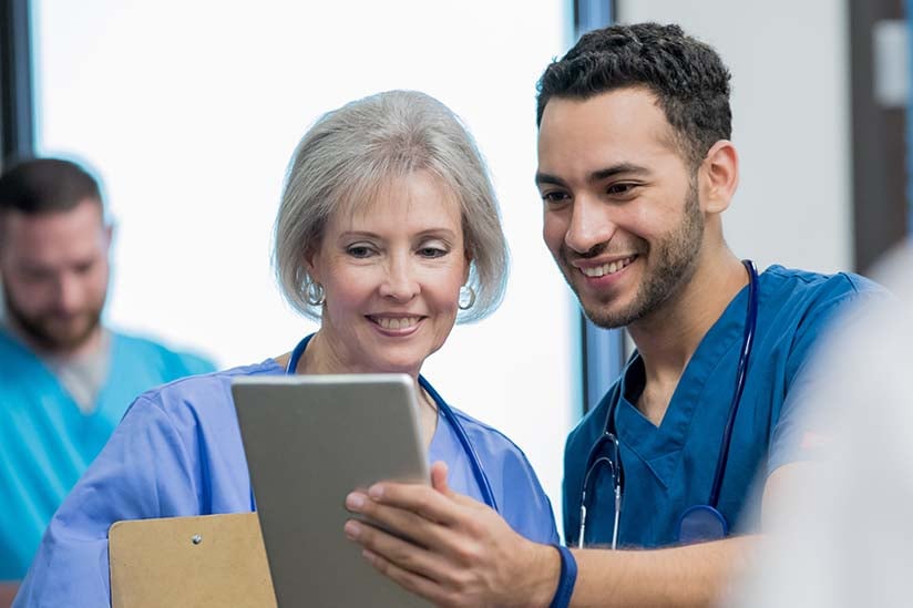 An older female nurse and a younger male nurse are standing together. The older nurse is reviewing some documentation that the younger nurse is holding in his hand and showing her. Both are smiling.