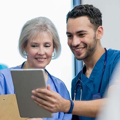 An older female nurse and a younger male nurse are standing together. The older nurse is reviewing some documentation that the younger nurse is holding in his hand and showing her. Both are smiling.