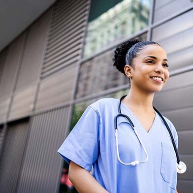 A smiling female nurse dressed in blue scrubs is walking outside of an office building.