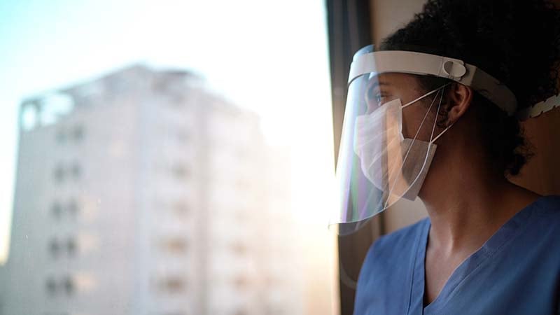 A nurse in blue scrubs and a face shield looks contemplatively out a window with a cityscape in the background, reflecting on the challenges faced during a healthcare crisis.