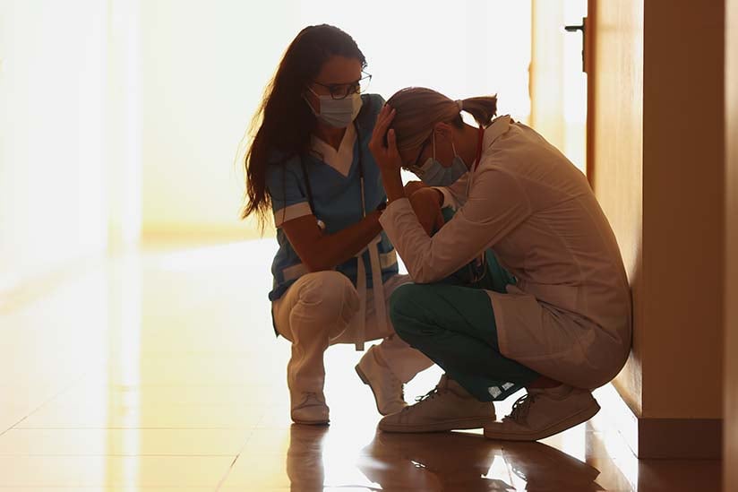 Two nurses in a corridor, one comforting the other who is crouched and appears distressed. They are both in scrubs and wearing masks, illuminated by warm light from the end of the hallway.