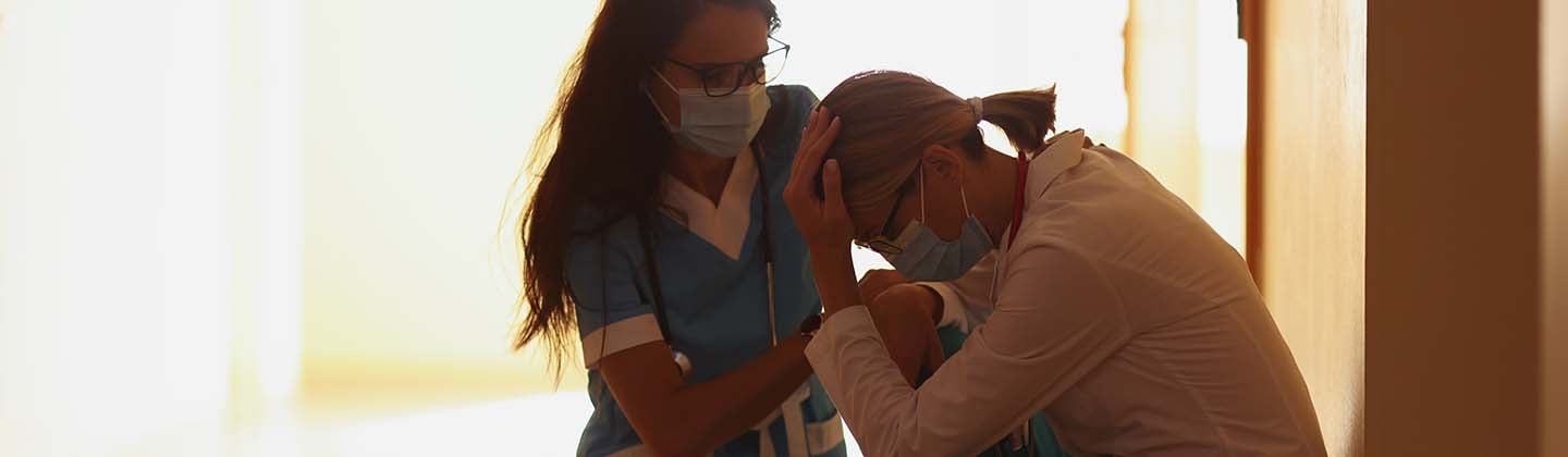 Two nurses in a corridor, one comforting the other who is crouched and appears distressed. They are both in scrubs and wearing masks, illuminated by warm light from the end of the hallway.
