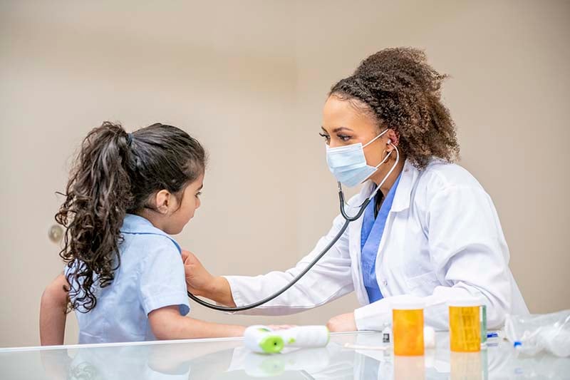 A pediatric nurse in a white coat and mask uses a stethoscope to listen to a young girl's heart, who looks at her with trust and curiosity during a medical check-up in a clinic.