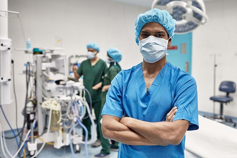 A confident nurse in blue scrubs and a surgical cap stands with arms crossed in an operating room, with medical equipment and colleagues working in the background.