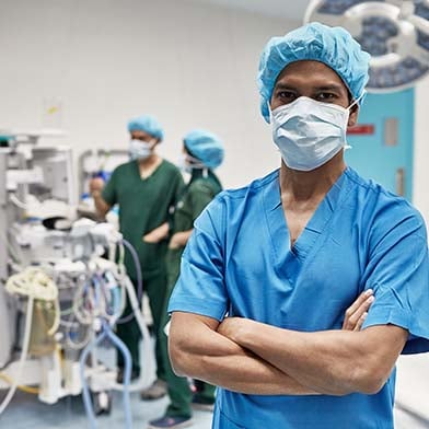 A confident nurse in blue scrubs and a surgical cap stands with arms crossed in an operating room, with medical equipment and colleagues working in the background.