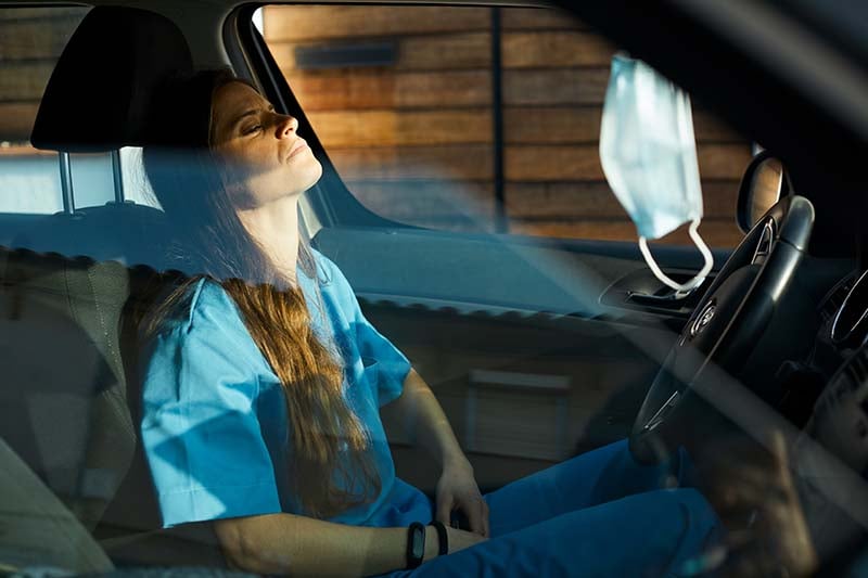 A female nurse is sitting in her parked car, her eyes are closed, and her head is back suggesting she is exhausted after a long shift.