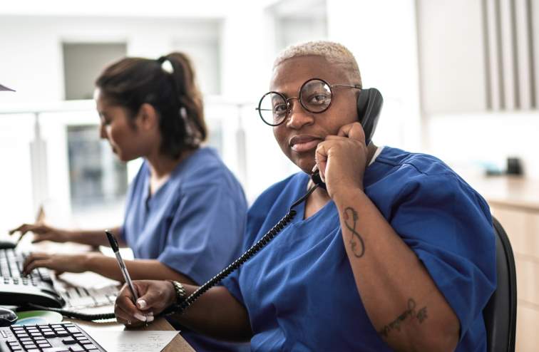 Black nurse wearing glasses speaking on the phone while sitting at a work station.