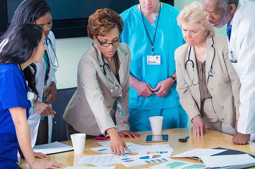 A group of healthcare professionals, including nurse administrators, are gathered around a table reviewing charts and graphs. There is a mix of attentiveness and discussion among the group, with some individuals pointing towards the documents. The setting suggests a collaborative meeting in a medical office environment.