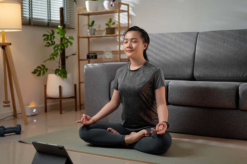 An Asian American nurse is sitting in a yoga position on a mat in a clam and serene looking room. She has her eyes closed in quiet mediation.