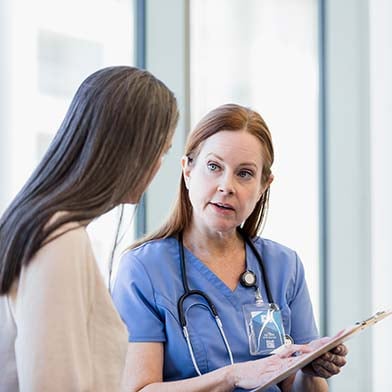 A nurse in blue scrubs with a stethoscope around her neck is attentively listening to a patient while holding a clipboard. They are standing in a brightly lit room with a large window in the background.