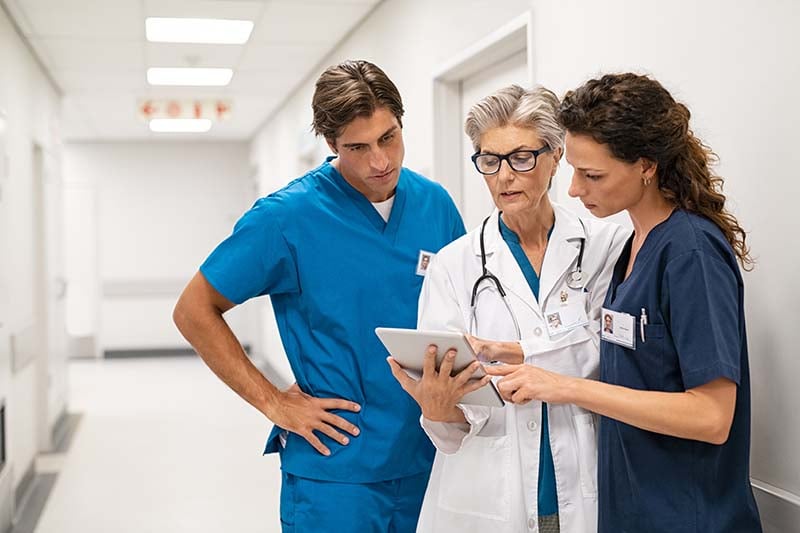 Three people are standing in a hospital corridor, a male nurse and two female nurses, and they are all looking intently at some information that one of the nurses is holding in her hands.
