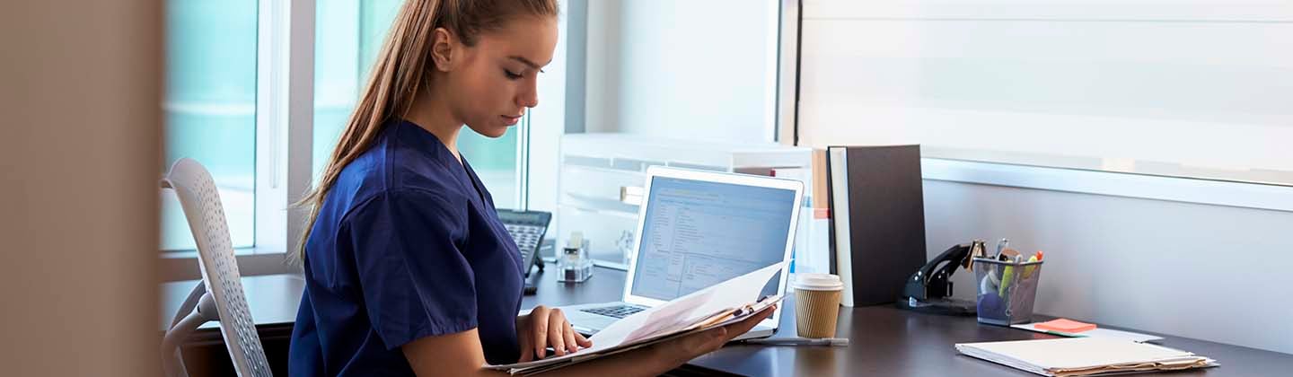 A female nurse wearing dark blue scrubs is seated at a desk and reading information from a folder. A computer screen can be seen in front of her.