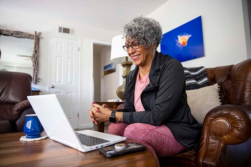 A smiling middle aged nursing student is seated at home, in front of her laptop, and actively participating in an online class.