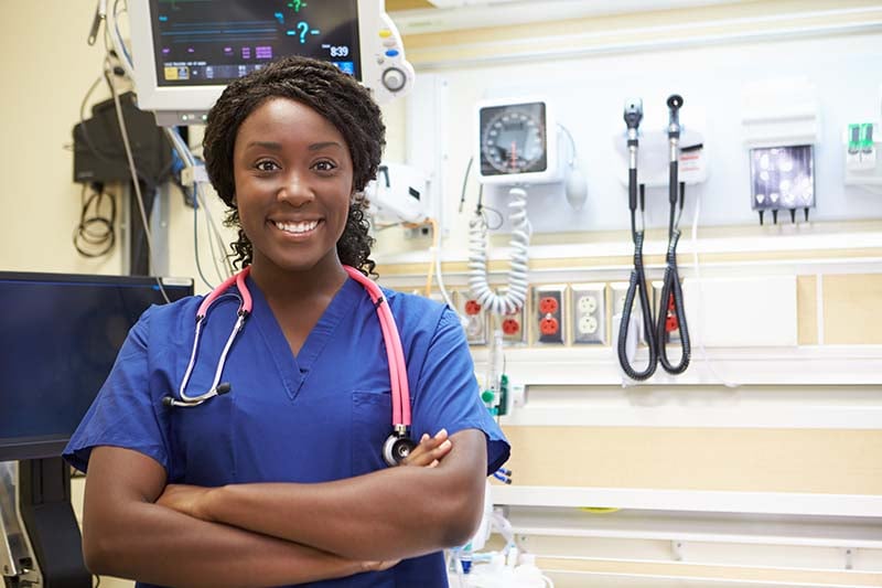 A confident nurse in navy blue scrubs with a pink stethoscope stands with crossed arms in an intensive care unit, surrounded by medical monitoring equipment, radiating professionalism and care.