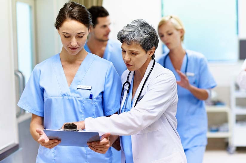 A group of nurses are in a hospital hallway walking and talking. An older nurse wearing a white lab coat is pointing to a clipboard a younger nurse is holding. She appears to be speaking about something that is documented on the clipboard while the younger nurse is listening. 