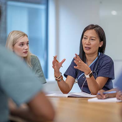 A female nursing leader is seated at a table and meeting with her staff, who are also seated. She is using her hands to emphasize a point, and her staff is looking at her and listening intently.