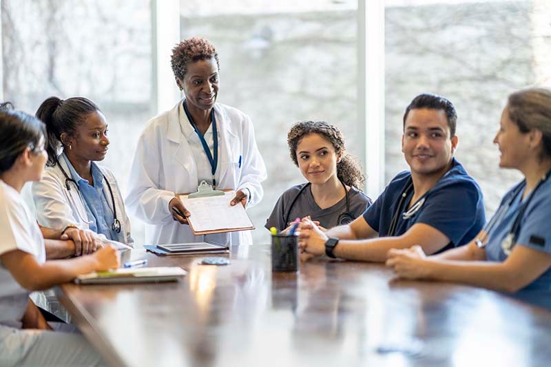 A female nursing leader is standing at a conference table, engaged in conversation with a group of her staff, who are seated around the table.