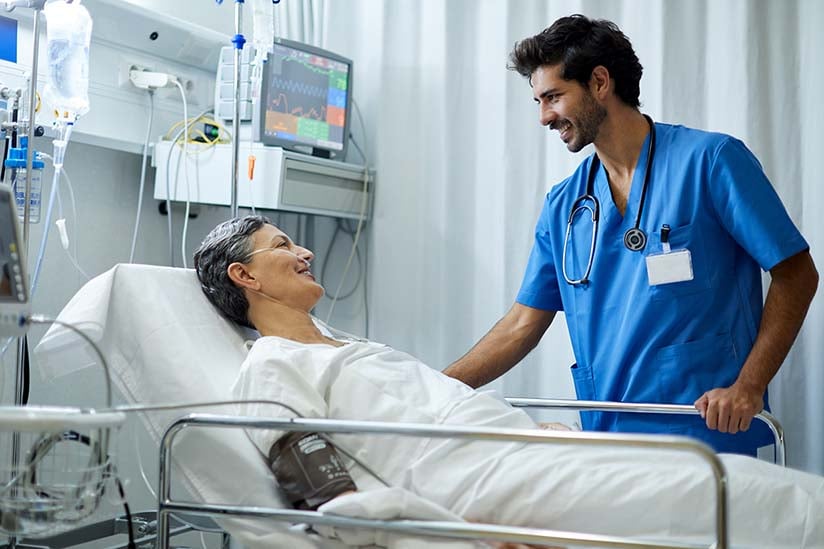 A male nurse is standing at the bedside of his hospitalized patient, smiling, and looking attentively. The patient is reclining and is attached to a blood pressure monitor and a nasal cannula.