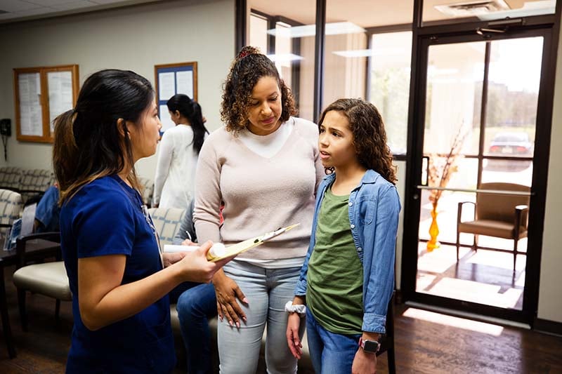 A female nurse is meeting with an adolescent patient and their mother in a medical center waiting room. The nurse is listening intently as the young patient is speaking to her.