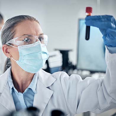 A female nurse is wearing a lab coat, protective eyewear, mask and gloves, and she is holding up and inspecting a blood specimen in a tube.