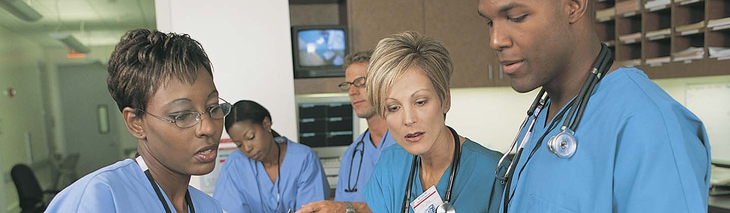Three nurses are standing at the nurse’s station in a hospital and having a discussion. There is printed information on the desk, and they are looking at information on a small screen, possibly a pager.