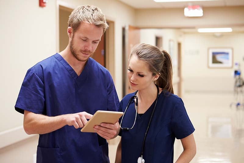 A male nurse and a female nurse are standing in the hallway of a hospital. They are looking at information on an electronic tablet in the male nurse’s hand.