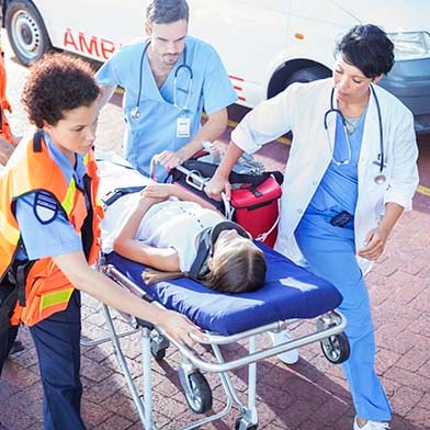Two Emergency Room staff members are helping a team of emergency medical technicians to push a stretcher into the Emergency Department. A young child wearing a neck brace is calmly laying on the stretcher.