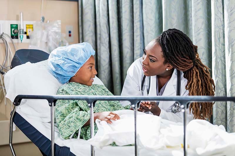 A beautiful African American nurse practitioner is seated next to a young child who is laying on a stretcher and wearing a hospital gown and a hair bonnet. The child is smiling and listening to the nurse practitioner speak.