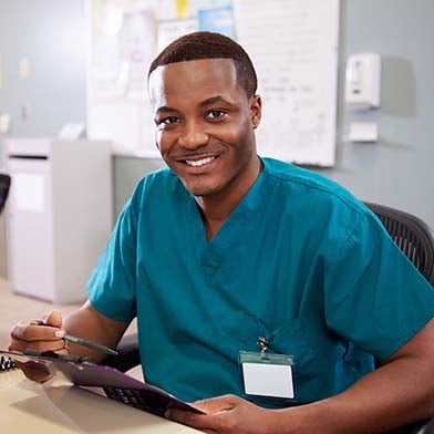 A smiling African American male nurse is seated at a computer workstation in a hospital ward. He is wearing blue scrubs and holding a clipboard and pen. 