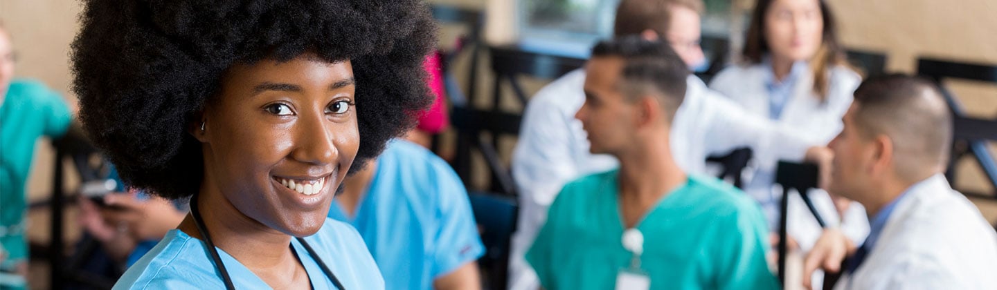 Smiling young nurse with an afro holds a clipboard in a classroom with fellow nurses chatting behind.
