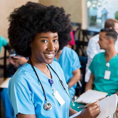Smiling young nurse with an afro holds a clipboard in a classroom with fellow nurses chatting behind.