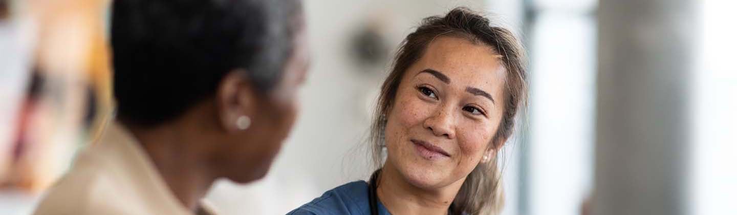 A cheerful nurse in blue scrubs takes notes on a clipboard while engaging with an elderly patient in a clinic room, signifying a caring and professional nurse-patient interaction.