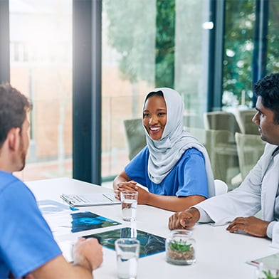 A group of culturally diverse nurses are seated at a conference table attending a meeting. They are smiling and engaged in conversation.