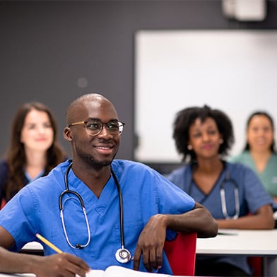 A diverse group of nursing students dressed in various color scrubs are engaged in learning in a classroom setting.